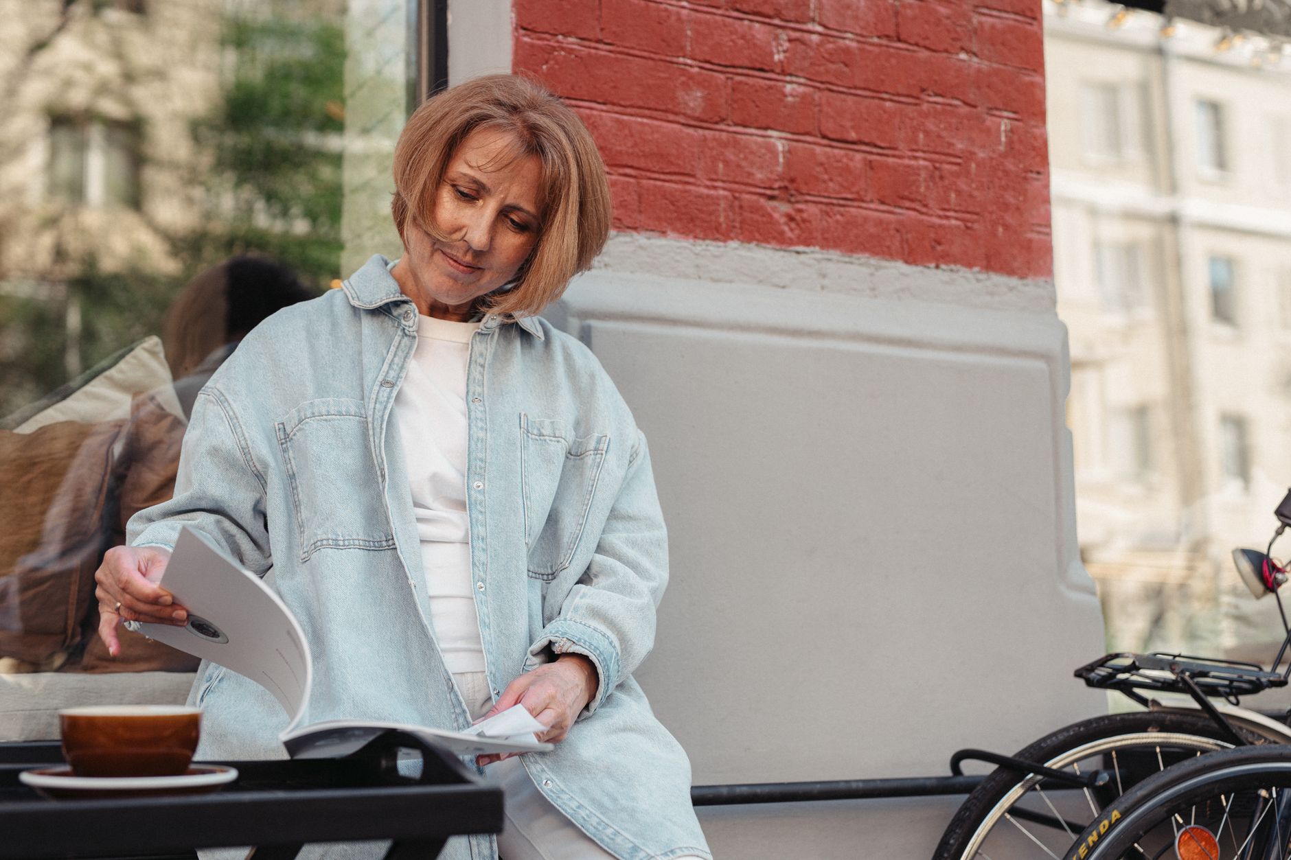 A woman in her fifties in a denim jacket reading a magazine beside a cup of coffee at an outdoor café table