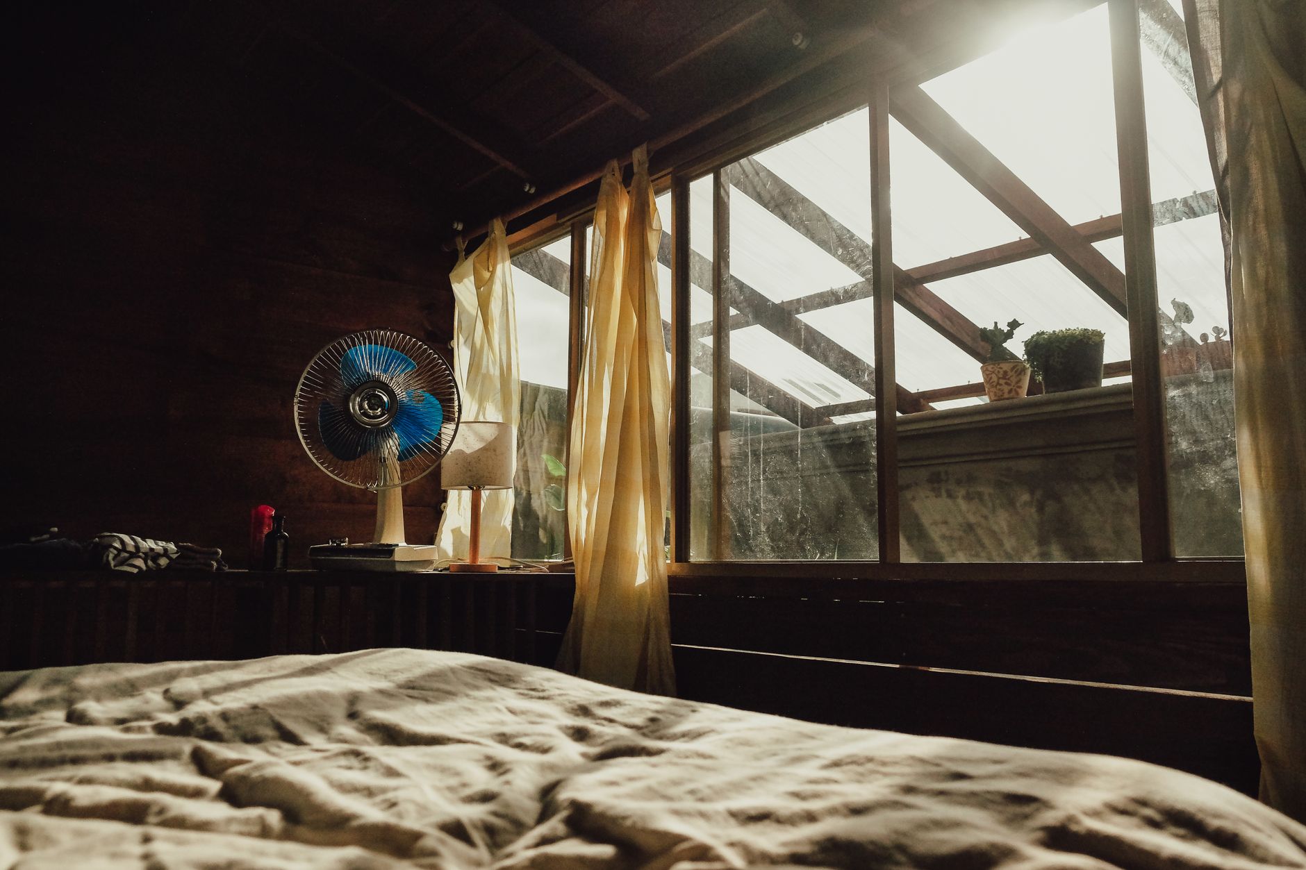 A desk fan beside an open window with sheer curtains, next to an unmade bed in warm morning light