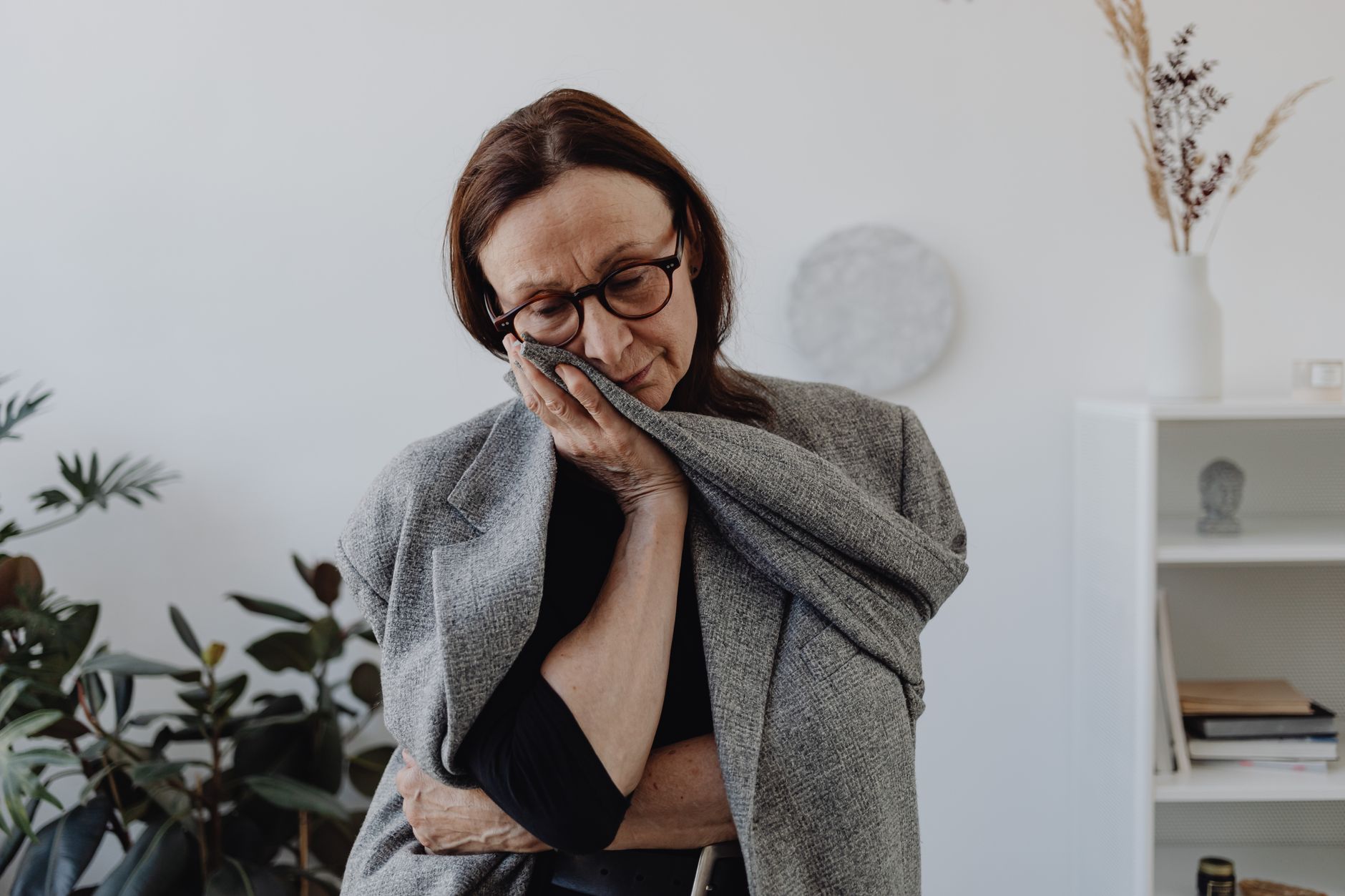 A woman in her fifties with glasses pressing the sleeve of a grey cardigan to her face, looking tired and unwell