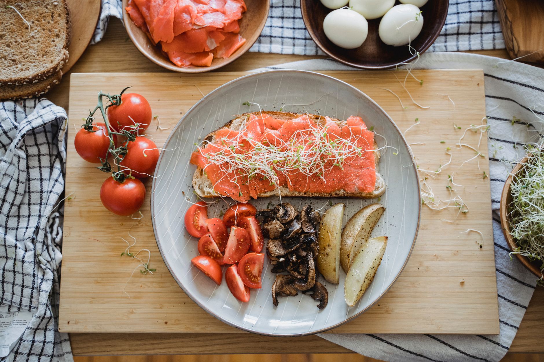 A plate of smoked salmon on rye toast with cherry tomatoes, mushrooms and potato wedges, eggs and bread on the table around it